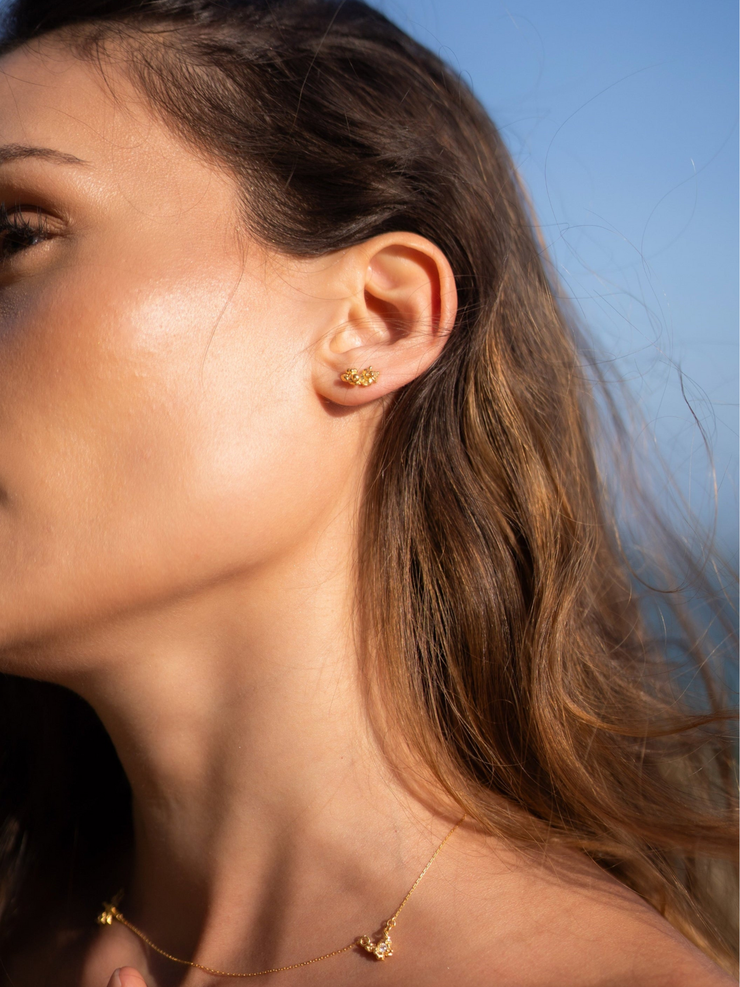 Close-up of a woman wearing gold earrings and a necklace against a blue sky.