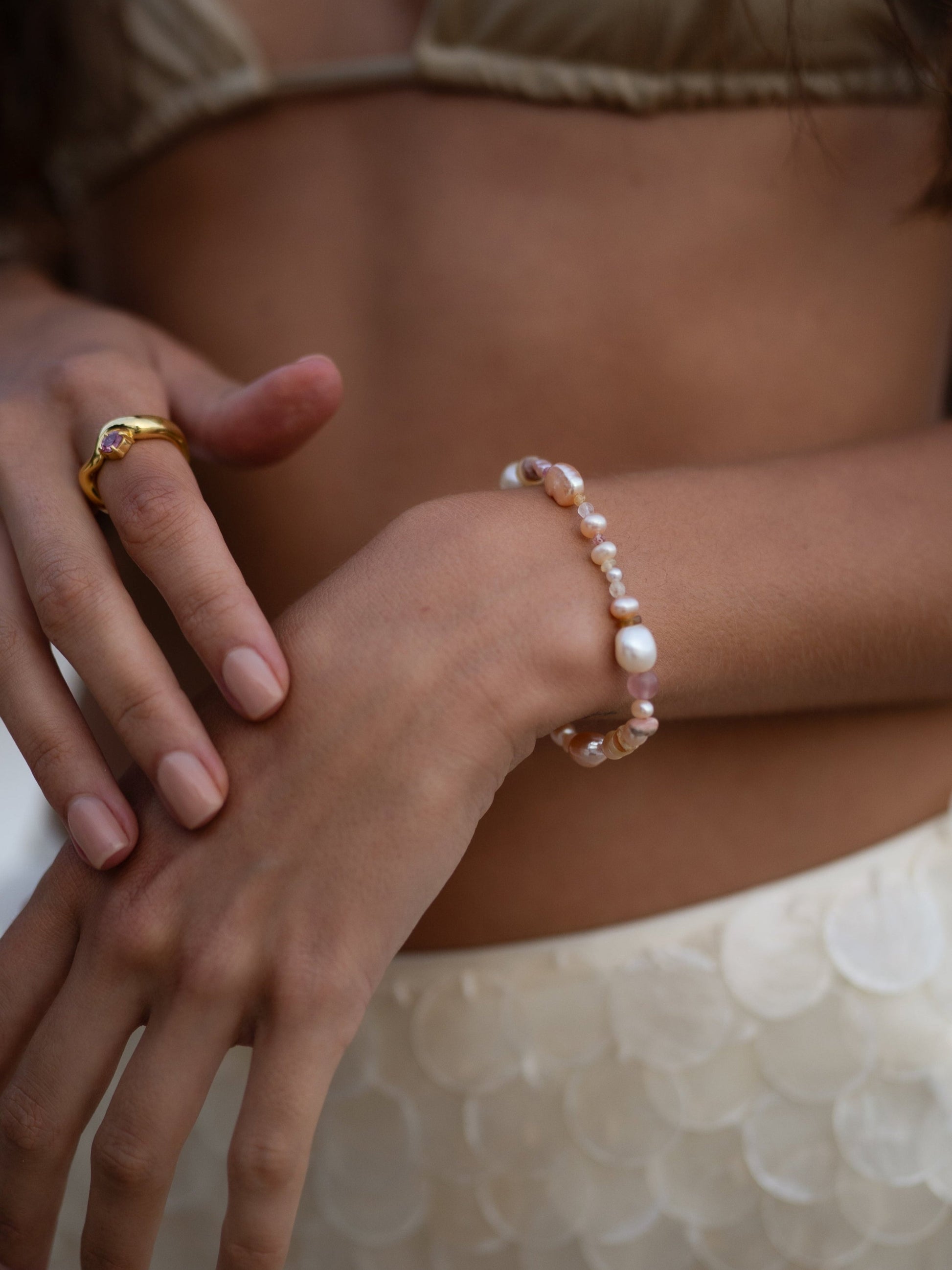 Close-up of a person's hands wearing a gold ring and pearl bracelet on a neutral background