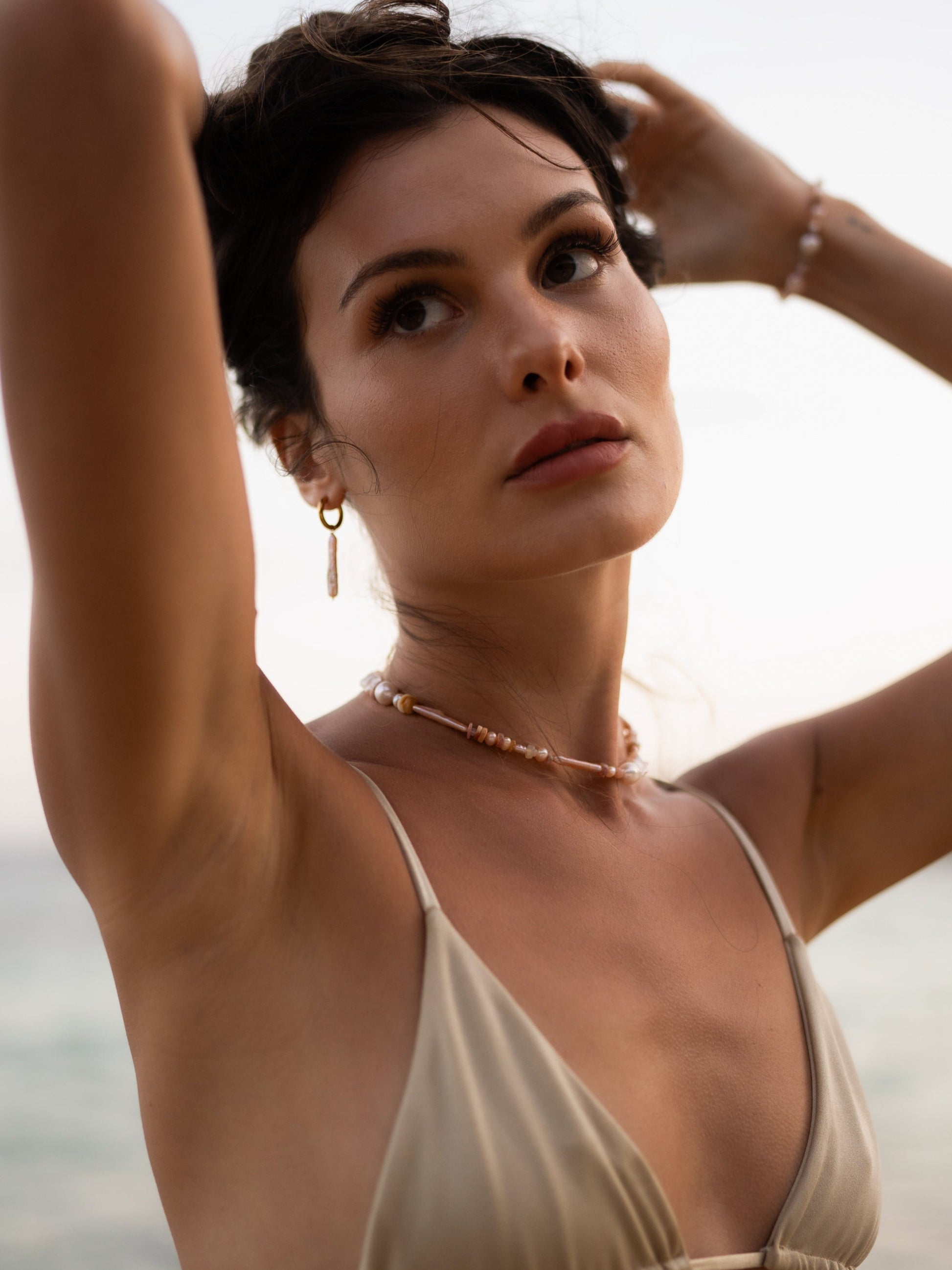 Woman in a beige bikini top with arms raised, wearing a necklace, against a blurred natural background.