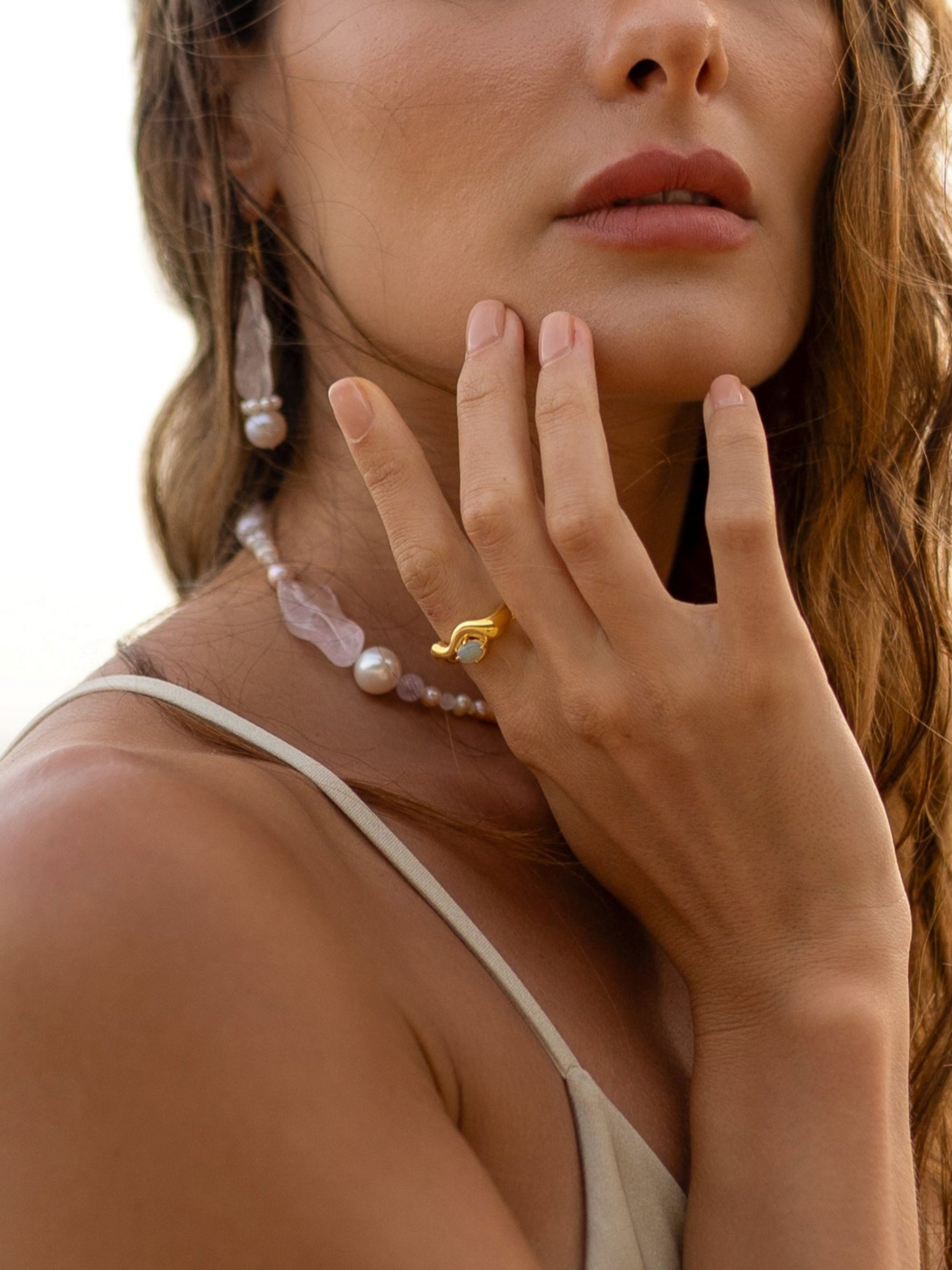 Close-up of a woman's hand wearing a gold ring with a pink gemstone, pearl necklace, and earrings against a blurred background.