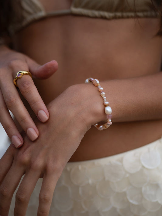 Close-up of a person's hands wearing a gold ring and pearl bracelet on a neutral background