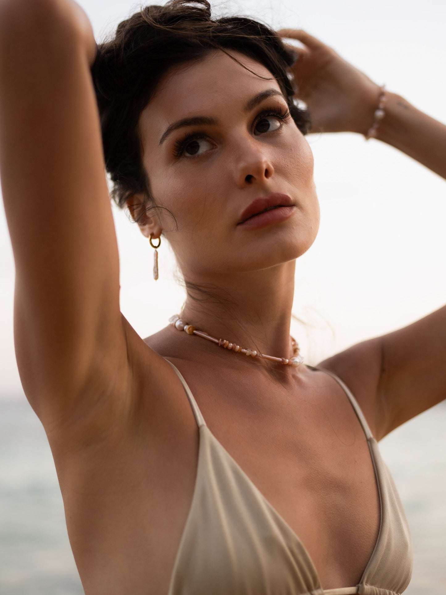 Woman in a beige bikini top with arms raised, wearing a necklace, against a blurred natural background.